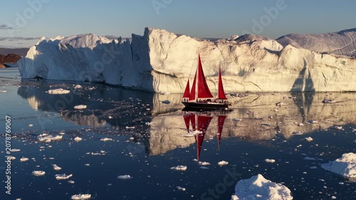 Sailboat with red sails cruises past giant arctic icebergs
