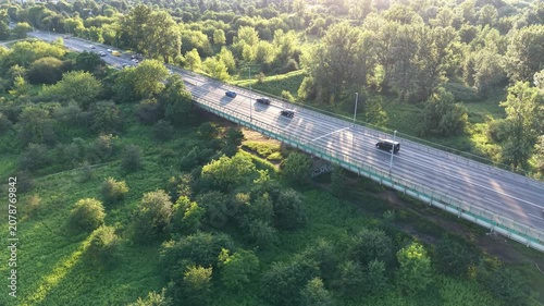 Aerial view of a winding road bordered by lush greenery and trees, showcasing vehicles traveling along the road with sunlight filtering through the foliage in a serene landscape