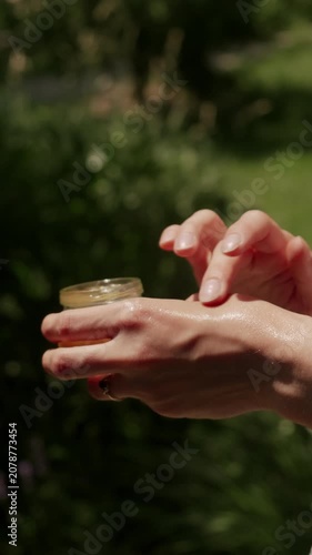 Closeup of adult hands poised over open cosmetic jar in sunlit light, with blurred green foliage backdrop