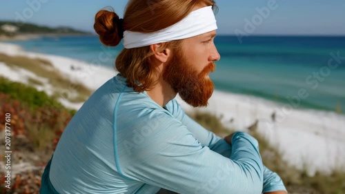 Red bearded man seated overlooking turquoise sea from dune