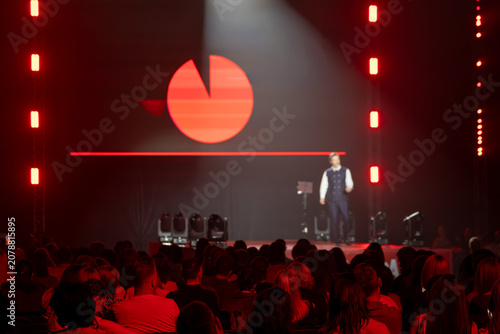 Business presenter on stage addresses seated audience under dramatic red lighting, logo projection visible on backdrop, conference atmosphere, public speaking, live event.