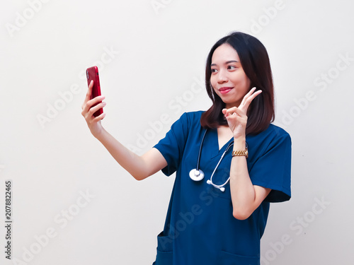 A cheerful Asian nurse in blue scrubs shows a peace sign while taking a selfie on her smartphone, representing modern clinical staff and healthcare lifestyle.