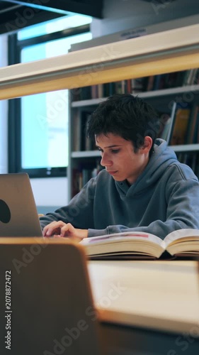 Vertical video of a focused teenage boy studying in a library, using a laptop and reading a book while preparing for exams.