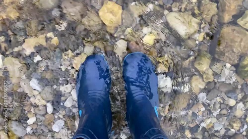 POV shot of legs in denim jeans and waterproof boots in flowing river water