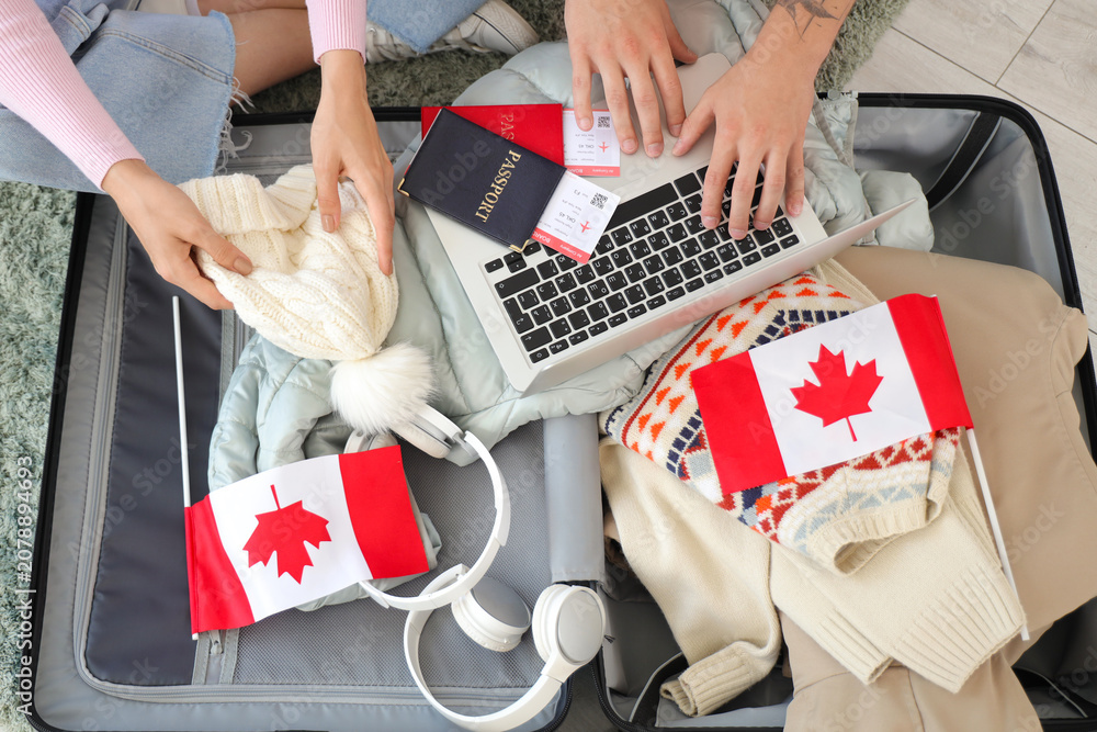 Obraz premium Young couple of tourists with Canadian flags and laptop packing suitcase at home, top view
