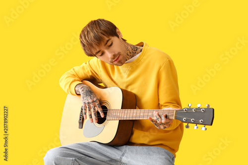 Cool tattooed young man playing guitar on yellow background
