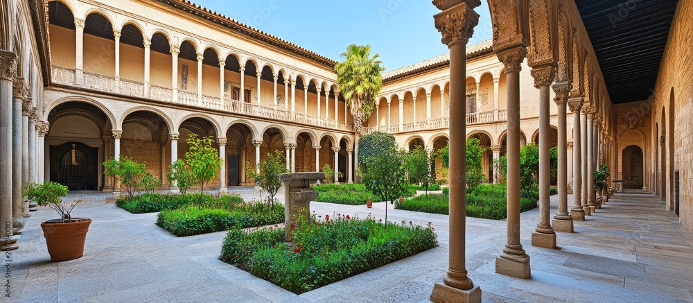 Fototapeta premium A serene courtyard with columns and arches surrounds a lush garden with trees and shrubs under a clear blue sky on a sunny day.