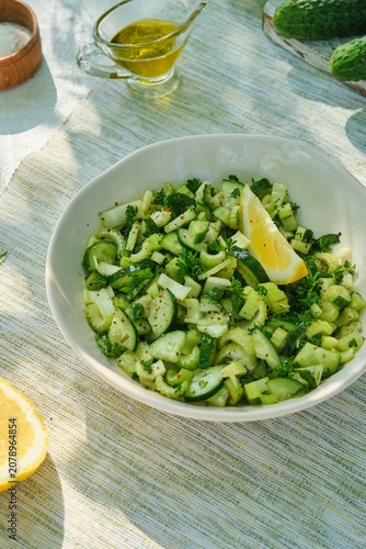 A healthy salad with celery, cucumber, parsley, and a lemon-olive oil dressing in a light bowl on a white concrete background.