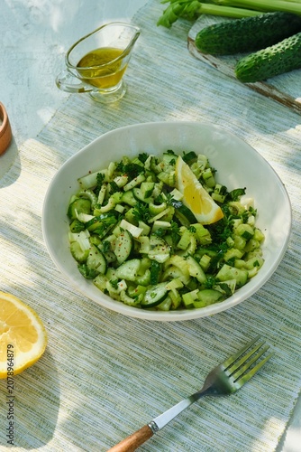 A healthy salad with celery, cucumber, parsley, and a lemon-olive oil dressing in a light bowl on a white concrete background.