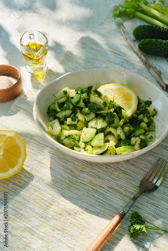 A healthy salad with celery, cucumber, parsley, and a lemon-olive oil dressing in a light bowl on a white concrete background.