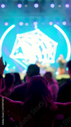 vertical video. A woman dances with her arms raised in front of a large stage at an awesome rock festival. A person dances in the crowd with an arm raised at a live concert. Blurred video.
