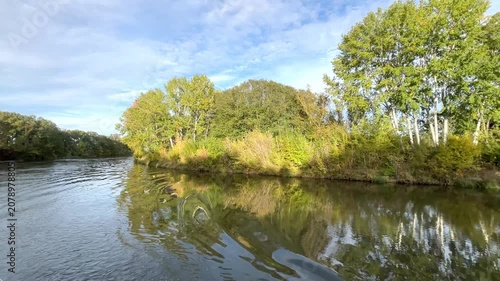 Calm river, yellow-green leaves on the trees, an idyllic river landscape seen from a boat in autumn, sunny day, blue sky, gentle waves, trees reflected in the river, green reflection