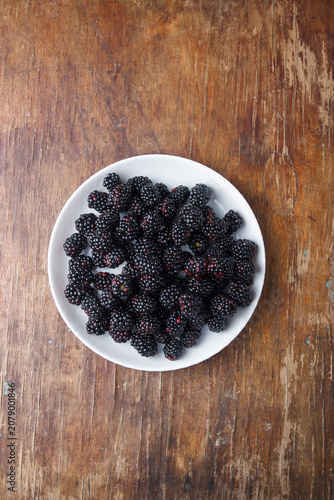 ripe fresh blackberries in white plate on brown old rustic wooden table, top view, copy space