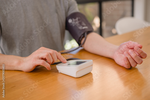 Man sitting at table while carefully using a digital blood pressure monitor on his arm to track his cardiovascular health and wellness at home during daylight