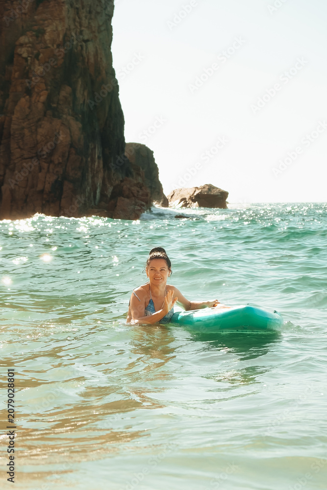 Obraz premium beach Ursa, Portugal. woman is in water holding surfboard while smiling. The ocean is bright and reflecting sunlight. Rocks are visible in the background near the shore.