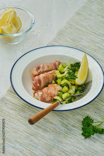Grilled cod wrapped in bacon strips with celery and cucumber salad on a white plate against a white concrete background.