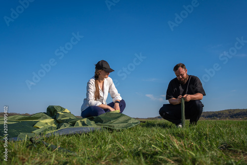 Young couple collaborating to unpack and assemble their green camping tent on a vast grassy field under a clear blue sky, preparing for an outdoor adventure