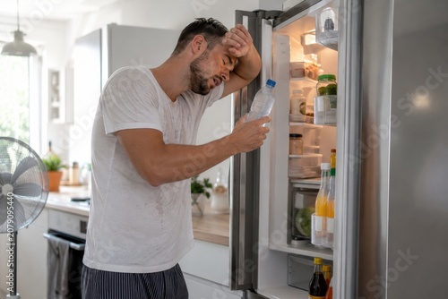 Young man feeling hot and tired while drinking water from the fridge in a modern kitchen