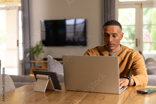 African American man typing on laptop at living room table, tablet and phone, wearing tan sweater