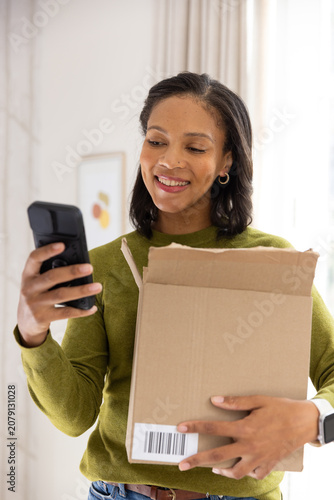 African American woman holding shipping box with barcode at chest checking phone at home