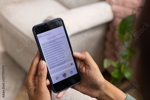 African American woman holding phone in navy case wearing green reading Business Proposal on sofa