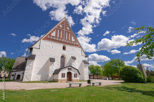 Historic Porvoo Cathedral stands beneath a dramatic blue sky with towering white clouds. The medieval Finnish church, with its whitewashed walls and separate bell tower, is a landmark of Porvoo's Old