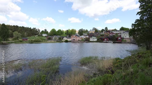 picturesque riverside of Porvoo, Finland. Colorful wooden houses line the far bank, nestled among lush green trees under a bright blue sky with fluffy white clouds. Reeds sway gently along the water's