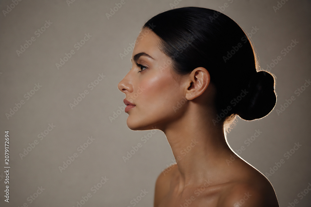 Obraz premium A woman with dark hair styled in a bun is shown in profile against a plain, neutral background.