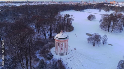 B-roll drone footage capturing the quiet winter morning at Munich's famous Monopteros landmark, with golden sunrise light illuminating the snow-covered public park meadows and sleeping trees.