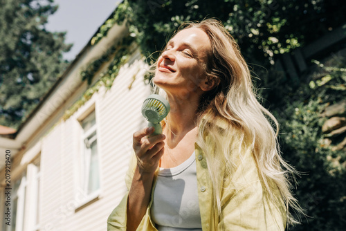 A happy smiling attractive blonde woman uses a portable electric fan outdoors on a hot midday.