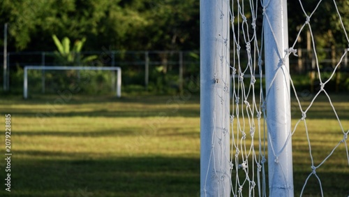 A selective-focus photo featuring an empty sports net, with the faint outline of practice goalposts visible in the distance against a rural backdrop.