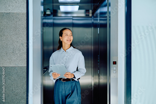 Cuadro en lienzo Young Asian businesswoman smiling, holding a digital tablet, and stepping out of