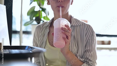 A young woman takes a sip from her drink while sitting at a table indoors. She is focused on her work in a vibrant environment with natural light coming through the windows