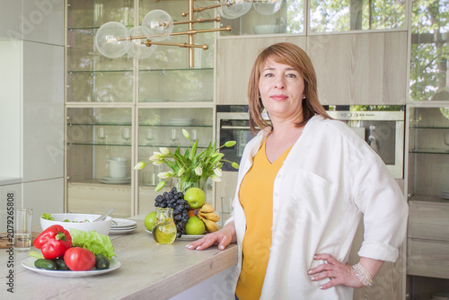 Woman stands in modern kitchen with fresh vegetables and fruits on table during sunny day