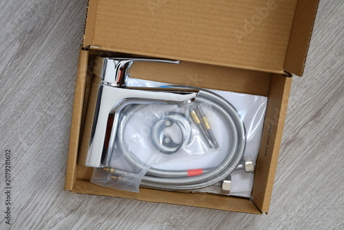 Top view of a new chrome water faucet mixer with installation kit inside an open cardboard box 