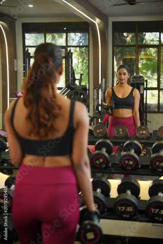 Obraz na płótnie Maharashtra, Sion, India- 22nd March 2026- Young Indian girl  in her active wear doing workout with dumbbells in gym, her reflection is seen in the mirror