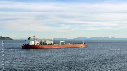 Ships are seen in a harbor transporting LNG. Various cargo vessels are docked, and the water reflects the surroundings. The location shows a busy port operation.