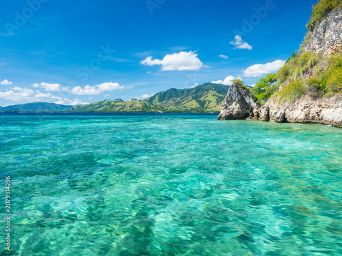Crystal clear turquoise waters surround a small tropical island in Riung 17 Islands National Park, Indonesia. A peaceful beach, lush green hills, and blue skies create a serene paradise