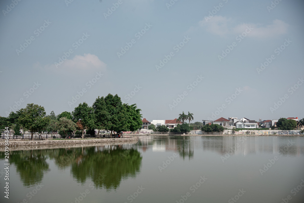 Fototapeta premium Scenic view of a calm urban lake surrounded by lush green trees and residential houses under a clear hazy sky.