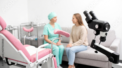 Professional female gynecologist consulting young patient in clinic room with pink gynecology chair and colposcope machine for health checkup exam
