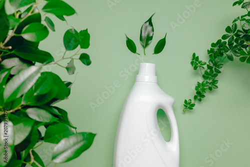 Mockup bottle laundry detergent or fabric softener close up with green plant leaves on green background top view. Natural washing and cleaning product for home.