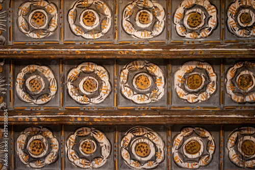 A section of an aged, ornate wooden ceiling adorned with a repeating pattern of painted Tudor roses, conveying a sense of historical artistry and enduring craftsmanship