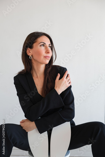 Portrait of young business woman looking side in black suit sits on white childs chair with bunny ears in white studio. Office fashion. Elegant style, fashion model. Flirting pose. High quality photo.