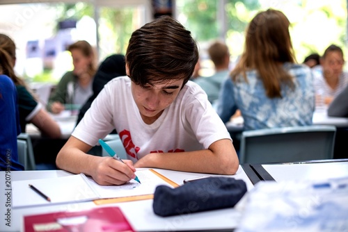 Fotografie Boy concentrating on writing at a classroom desk with peers around