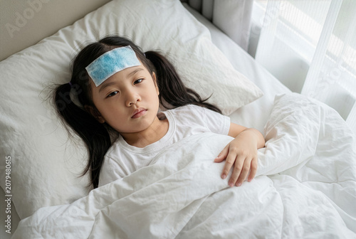Sick little Asian girl lying in bed with a cooling gel patch on her forehead, suffering from high fever, cold or flu.
