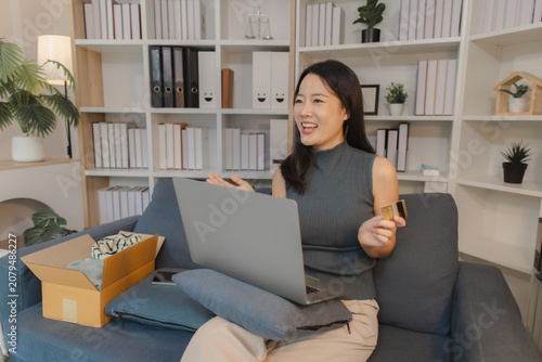 Young Asian woman using laptop and holding credit card at home, smiling while making online payment, representing ecommerce activity, digital transaction, and convenient modern cashless lifestyle.