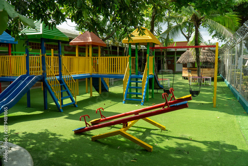 Фотография Colorful empty outdoor kids playground with slides, swings, see-saw and artificial green grass turf on a sunny day surrounded by trees
