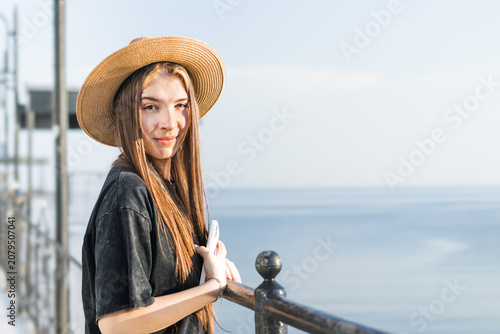 Young woman with long brown hair wearing a straw hat leans on a railing by the sea, with calm water and a clear sky in the background