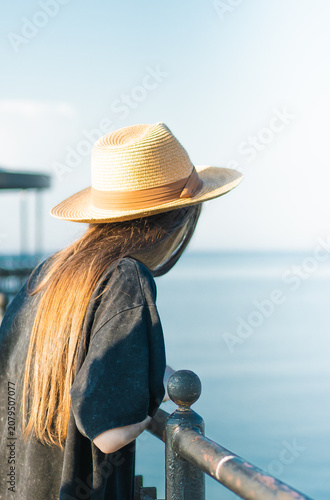 Young woman with long hair wearing a straw hat leans on a railing, gazing at the calm sea under a clear blue sky during a sunny day