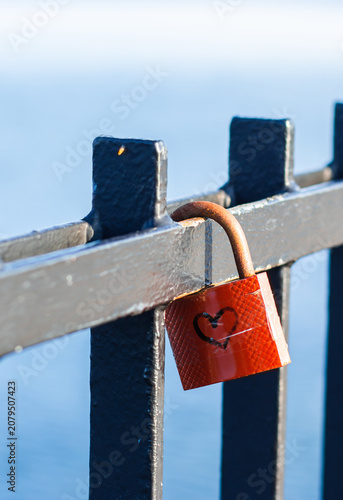 Red padlock with heart symbol hanging on a black metal fence against a blurred blue background of water and sky